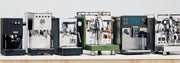 A lineup of six professional espresso machines in various colors including black, silver, green, and white, arranged on a light wooden counter against a plain light gray background.