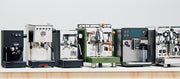 A lineup of six professional espresso machines in various colors (black, silver, green, and stainless steel) arranged on a light wooden counter against a plain white background.