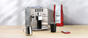 Close-up of a Gaggia Brera in Silver next to a bag of Gaggia Milano Intenso Coffee a cup, cleaning tablets and a milk frothing pitcher.