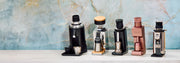Five countertop coffee grinding devices displayed side by side on a white surface against a light blue marble-patterned background. The products include black, silver, gold, and rose gold color options.