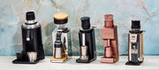 Five countertop coffee grinding devices displayed side by side on a white surface against a light blue marble-patterned background. The products include black, silver, gold, and rose gold color options.