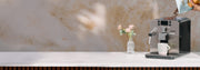 Person pouring Whole Latte Love Crema Wave coffee beans into a silver Gaggia Brera Espresso Machine on a white counter next to pink flowers in front of a brown wall. 