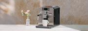 Close-up of a silver Gaggia Brera Espresso Machine on a white counter next to pink flowers in front of a brown wall. 