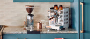Close-up of a Rocket Appartamento and Rocket Grinder on a marble kitchen counter next to a stove and other espresso brewing accessories.