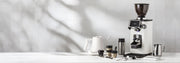 Ceado REV Zero grinder in white next to a varia smart kettle, notNeutral pico cups, a revolution milk frothing pitcher, spoons, scale and tampers on a white counter top in front of a white tiled wall.