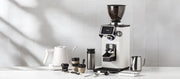 Close-up of a Ceado REV Zero grinder in white next to a varia smart kettle, notNeutral pico cups, a revolution milk frothing pitcher, spoons, scale and tampers on a white counter top in front of a white tiled wall.