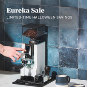 Persons hand pressing grind options on a Eureka Oro Mignon XL in white on a white counter with blue tiles behind it. Promotional text overlayed that reads "Eureka Sale - Limited-Time Halloween Savings".