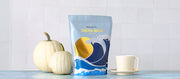 Close-up of Whole Latte Love Crema Wave on a blue counter next to a coffee mug and white pumpkins in front of a white tiled wall. 