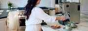 Woman in white brewing coffee from a Gaggia Classic GT on a marble kitchen island with 2 sage notNeutral pico coffee cups next to it.