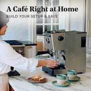 Close-up of a woman in white brewing coffee from a Gaggia Classic GT on a marble kitchen island with 2 sage notNeutral pico coffee cups next to it. Promotional text overlayed that reads "A Cafe Right at Home - Build Your Setup & Save".