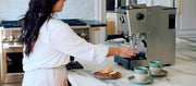Close-up of a woman in white brewing coffee from a Gaggia Classic GT on a marble kitchen island with 2 sage notNeutral pico coffee cups next to it.