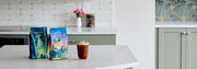 A bag of Whole latte Love Beaned UP and Whole Latte Love Buzzopolis next to a cup of coffee on a white kitchen island.