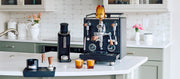 Close-up of a Profitec DRIVE Espresso Machine in black with wooden accents next to espresso accessories on a kitchen counter. 