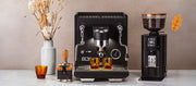 Close-up of a ECM Estetika Espresso Machine in black next to an ECM Exacto Grinder in black a Wiedemann 2.0 tamping station a tamper and fall foliage in a vase on a white counter top in front of a brown wall.