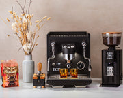 ECM Estetika Espresso Machine in black next to an ECM Exacto Grinder in black a Wiedemann 2.0 tamping station a tamper and fall foliage in a vase on a white counter top in front of a brown wall.