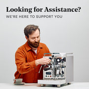 A man in an orange shirt is operating a stainless steel espresso machine at a table, with a coffee cup nearby. Large text at the top reads, 'Looking for Assistance? WE'RE HERE TO SUPPORT YOU.