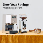 Gaggia Classic E24 next to a Eureka Silenzio 55 on a golden fabric next to an espresso martini and a disco ball with promotional text that reads "New Year Savings".