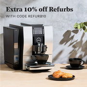 JURA coffee machine on a wooden table with two black cups and a plate of pastries, against a gray wall with promotional text that reads "Extra 10% off Refurbs with code REFURB10".