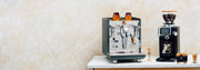 ECM Synchronika Espresso Machine in Anthracite next to a Ceado REV grinder and other Wiedemann espresso tools on a white counter in front of a brown and white wall.