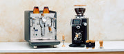Close-up of an ECM Synchronika Espresso Machine in Anthracite next to a Ceado REV grinder and other Wiedemann espresso tools on a white counter in front of a brown and white wall.