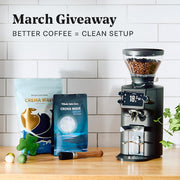 Close-up of the Mahlkonig E64 WS Espresso grinder and coffee on a wooden surface with a white tiled wall background and promotional text that reads "March Giveaway - Better Coffee = Clean Setup".