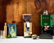 Gaggia Classic E24 in Green next to a Eureka Silenzio 55 in Gold on a white counter in front of a wooden background.
