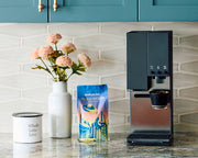 xBloom coffee maker next to a bag of Whole Latte Love Buzzopolis coffee, flowers and whole latte love branded jar on a kitchen counter.