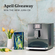 Jura C9 espresso machine next to package of Whole Latte Love Beaned UP and flowers on a marble background, promoting an April Giveaway.