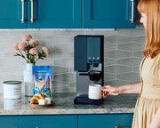 Woman making coffee using an xBloom coffee maker next to a bag of Whole Latte Love Buzzopolis coffee on a counter in a kitchen with teal cabinets and a gray backsplash.