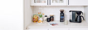 Bonavita Coffee Maker next to a Baratza Grinder, coffee canisters, coffee bag and cups in a white kitchen coffee nook.