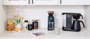 Close-up of a Bonavita Coffee Maker next to a Baratza Grinder, coffee canisters, coffee bag and cups in a white kitchen coffee nook.