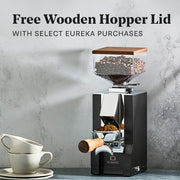 Eureka Mignon Silenzio 55 with Walnut accents on a gray stone counter and gray background next to latte cups. Promotional text overlayed that reads "Free Wooden Hopper Lid with Select Eureka Purchases".