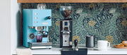 Close-up of a kitchen setup with Profitec GO Espresso Machine in blue, Eureka grinder, cups, tamper, coffee bags on a shelf and decorative elements in a coffee nook.