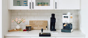 Close-up of a Gaggia Classic E24 in white with tiger maple accents next to a Varia VS4 and other coffee accessories in a white kitchen setting and coffee nook.