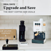 Gaggia Classic E24 in white with tiger maple accents next to a Varia VS4 and other coffee accessories in a white kitchen setting and coffee nook. Promotional text overlayed that reads "Deal Days: Upgrade and Save - The best coffee bar deals".