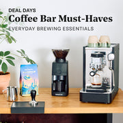 Stone Lite espresso machine next to a Baratza grinder and Whole Latte Love Buzzopolis coffee on a wooden counter top. Promotional text overlayed that reads "Deal Days: Coffee bar Must-Haves - Everyday brewing essentials".