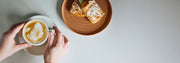 Person holding a cup of coffee with latte art next to a plate of pastries on a light surface.