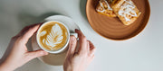 Close-up of the person holding a cup of coffee with latte art next to a plate of pastries on a light surface.