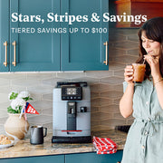 Woman drinking coffee in a kitchen next to a Gaggia Cadorna in Moonlight Gray on a brown granite counter next to flowers. Promotional text overlayed that reads "Stars, Stripes & Savings - Tiered Savings up to $100".