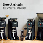 Row of coffee grinders with text 'New Arrivals: The Latest in Brewing!' on a tiled wall background.