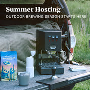 Gaggia Classic Pro E24 in Black with walnut accents outside on a truck tailgate next to a Varia hand grinder in white, tamper, mug and bag of Whole Latte Love Buzzopolis. Promotional text overlayed that reads "Summer Hosting - Outdoor brewing season starts here".