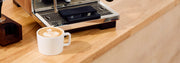A white ceramic cup of latte with leaf latte art sits on a light wood countertop next to a stainless steel espresso machine.