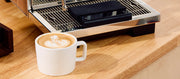 A white ceramic coffee mug filled with latte art sits on a wooden countertop in front of a stainless steel espresso machine.