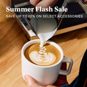 A person pouring milk from a metal pitcher into a white ceramic cup of latte art coffee on a wooden table, with promotional text reading 'Summer Flash Sale' at the top.