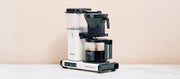 Close-up of the Technivorm Moccamaster in white on a white counter in front of a light pink background.