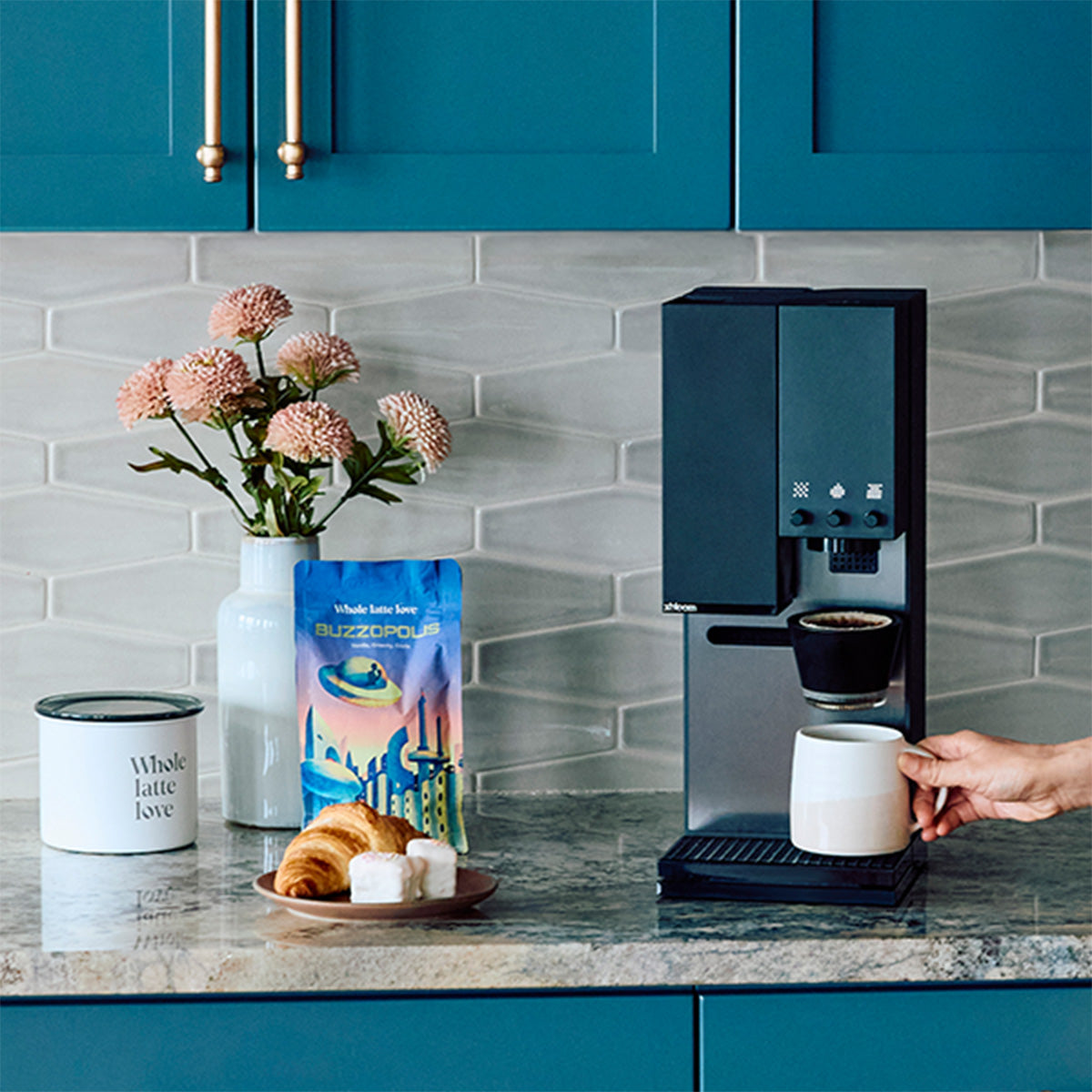Close-up of a person grabbing a coffee from the xBloom Studio in Black on a kitchen counter with blue cupboards next to  Whole Latte Love Buzzopolis, coffee canister, pastry and flowers.