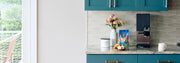 xBloom Studio in Black on a kitchen counter with blue cupboards next to  Whole Latte Love Buzzopolis, coffee canister, pastry and flowers.