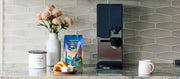 Close-up of a xBloom Studio in Black on a kitchen counter with blue cupboards next to  Whole Latte Love Buzzopolis, coffee canister, pastry and flowers.