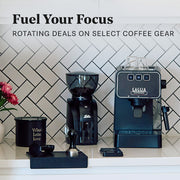 Gaggia Espresso Evolution in Black next to a Solis Grinder and other coffee accessories in front of a tile wall and on a white counter top. Promotional text overlayed that reads "Fuel Your Focus - Rotating Deals on Select Coffee Gear".