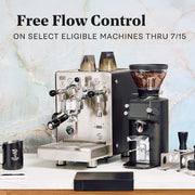 Promotional product photo showing a stainless steel espresso machine and a black coffee grinder on a countertop, with text reading 'Free Flow Control ON SELECT ELIGIBLE MACHINES THRU 7/15' above them. A marble-pattern backdrop and a few small accessories are visible around the machines.